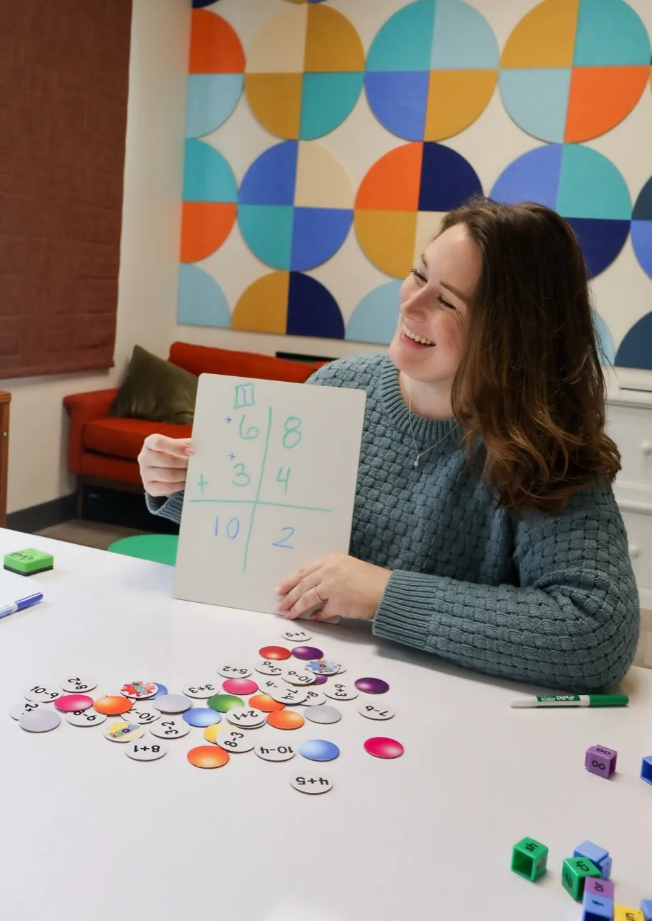 An educational therapist smiling while holding up a whiteboard with a math problem during a session.